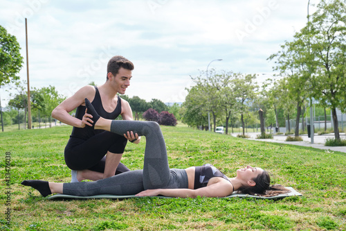 Personal trainer helping young woman in a park with stretching exercises. Leg stretch laying on a mat outside.