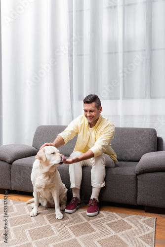 labrador dog sticking out tongue near cheerful man sitting on couch at home