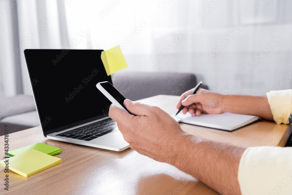 Fototapeta premium cropped view of freelancer writing in notebook while holding smartphone near laptop with blank screen