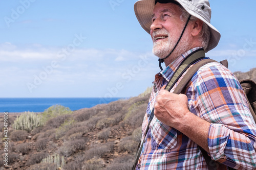 Smiling senior man with backpack and hat in outdoor trekking in arid landscape with sea on background