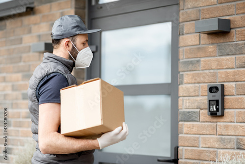 Tableau sur toile Courier in cap wearing mask and gloves holds a parcel box and staying near front door of house