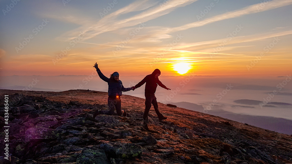 A couple holding hands and jumping on top of Babia Gora, Poland, with the panoramic view on sun rising above the horizon. Thick clouds below. The sky is pink and orange. Love and passion