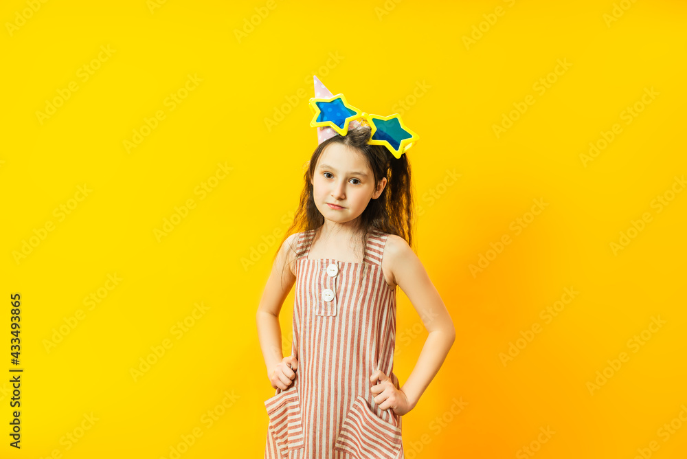 Portrait of young little brunette girl on yellow studio background in red striped dress and festive cap. Birthday. Frustrated facial expression. Portrait of young little brunette girl on yellow studio background in red striped dress and festive cap. Birthday. Frustrated facial expression.