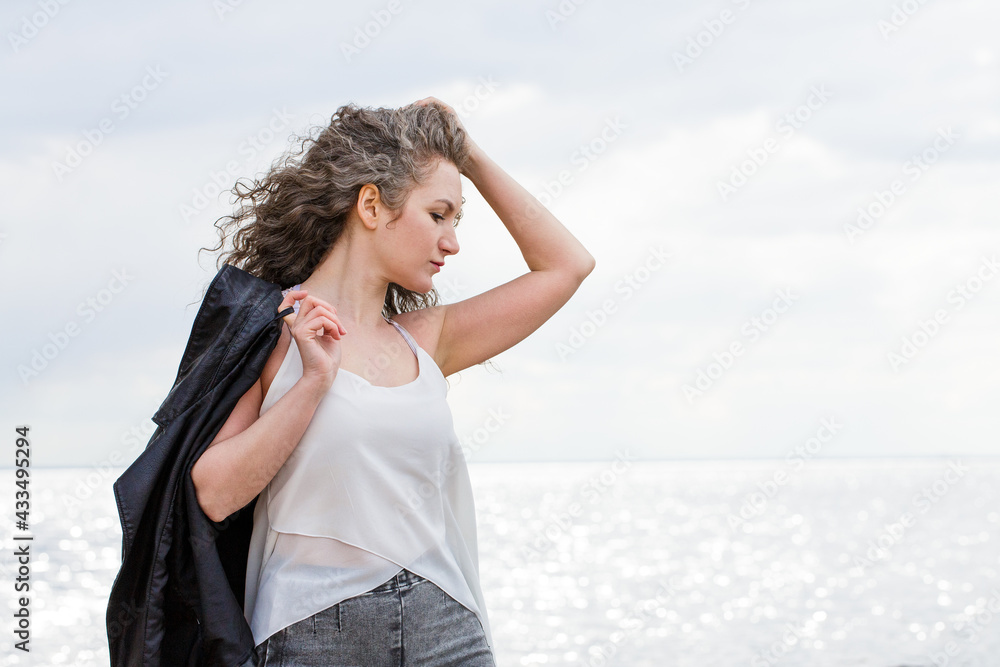 Portrait of a caucasian sad curly woman in a white blouse and black leather jacket standing on the seashore in the wind straightens her hair Portrait of a caucasian sad curly woman in a white blouse and black leather jacket standing on the seashore in the wind straightens her hair
