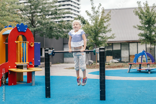 Happy smiling young girl exercising on children pull-ups bar outdoor at playground park. Child hanging on high bar. Summer fun activity workout and fitness. Kids sports and recreation outside.