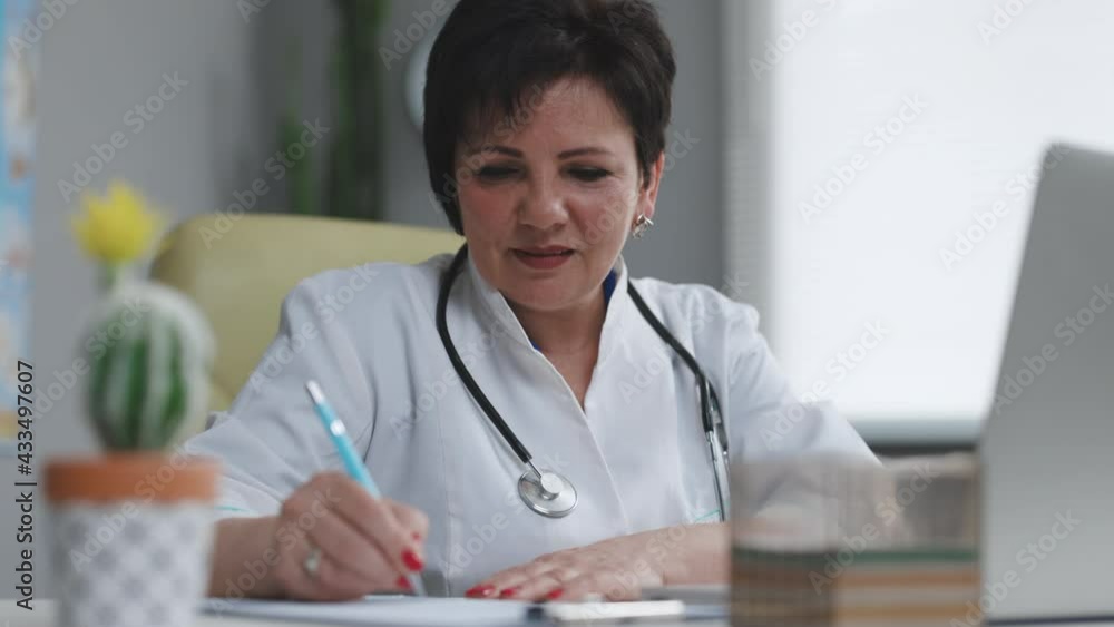 Middle aged senior head doctor in white medical coat sitting at workplace, talking to patient making video call on laptop, writing notes in paper journal Middle aged senior head doctor in white medical coat sitting at workplace, talking to patient making video call on laptop, writing notes in paper journal