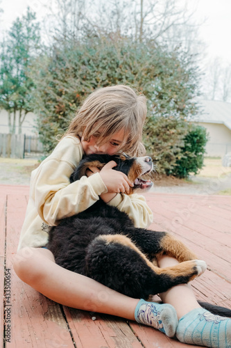 Adorable cute child girl sitting with domestic dogs and hugging little Bernese puppy on the porch of the house. Dogs. Concept of love to animals and friendship with pets.