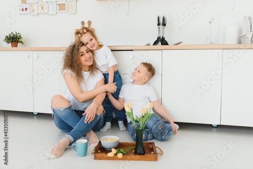 Happy young family, mom with little children sit on warm wooden floor in new modern design kitchen, mother with excited small kids relax rest in own renovated apartment, moving concept