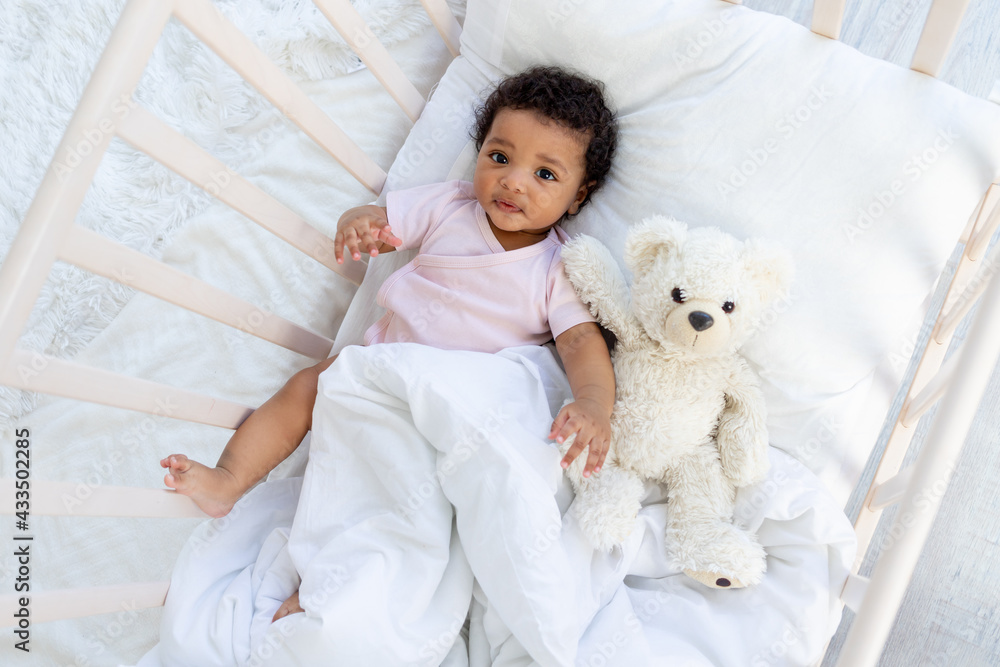 happy smiling african american baby in a crib with a teddy bear falls asleep or goes to bed
