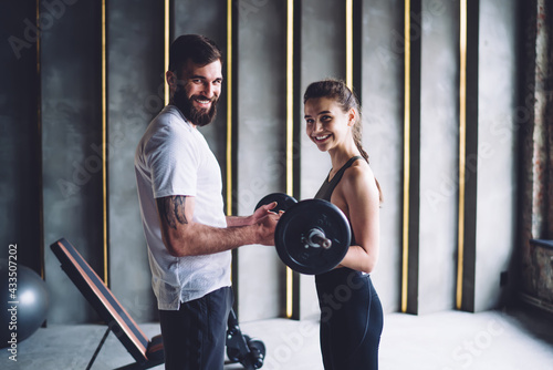 Half length portrait of cheerful young male and female friends in active wear satisfine with workout in gym, man fitness instructor helping happy woman client keeping perfect figure on workout