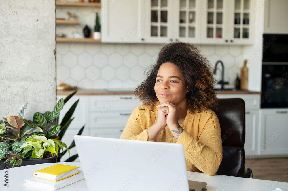 Delighted African-American young woman sits with a laptop in the kitchen in cozy apartment, looks away and dreaming, thoughtful happy dark skinned female freelancer lost in dreams and imaginations Delighted African-American young woman sits with a laptop in the kitchen in cozy apartment, looks away and dreaming, thoughtful happy dark skinned female freelancer lost in dreams and imaginations