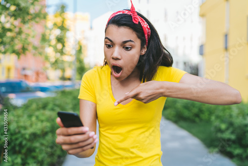 Outdoor portrait of young african american woman pointing with index finger at her modile phone screen with shocked face expression, astonished by her victory in online lottery