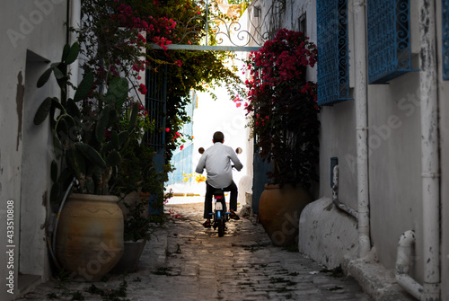 Motorcyclist rides down a narrow street. View of the old cute street with flowers. Retro italian moped.