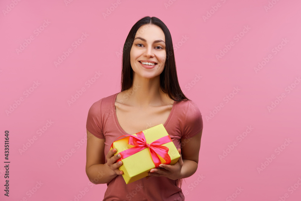 Fototapeta premium Portrait of happy attractive young brunette woman holding gift box, isolated over pastel pink background