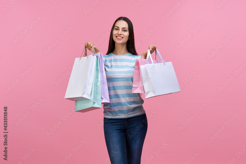  Studio shot of casually dressed beautiful smiling brunette girl posing with bunches of paper shopping bags in hands, isolated over pastel pink background
