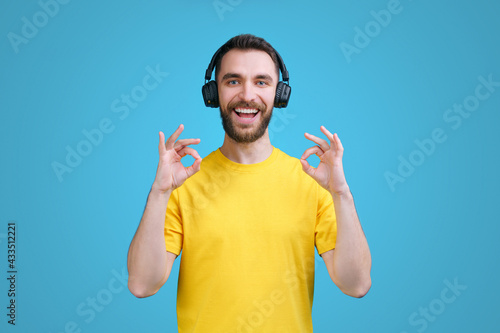 Portrait of cheerful young man in yellow t-shirt and wireless headphones showing ok sign gesture with his hands, isolated in blue background
