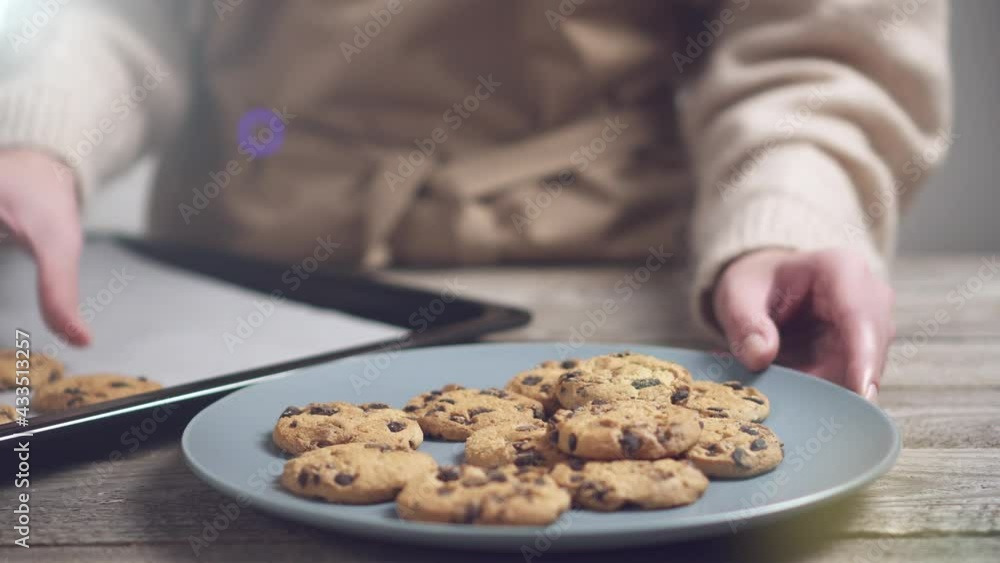 Homemade chocolate chip cookies on a baking sheet lined with parchment paper. A girl in a beige sweater puts cookies on a white plate. A slide of chocolate chip cookies on a wooden kitchen table. Homemade chocolate chip cookies on a baking sheet lined with parchment paper. A girl in a beige sweater puts cookies on a white plate. A slide of chocolate chip cookies on a wooden kitchen table.