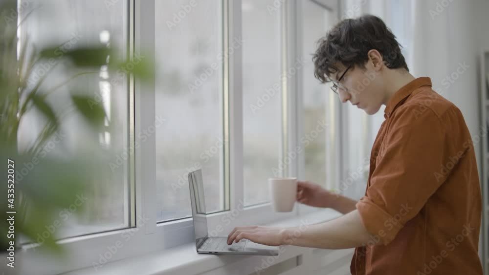 Busy young man working on laptop near window, freelancer working on project Busy young man working on laptop near window, freelancer working on project