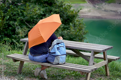 Rear view of a man with an open umbrella and backpack who sitting at a wooden picnic table on a green lawn with trees and a lake in the background.