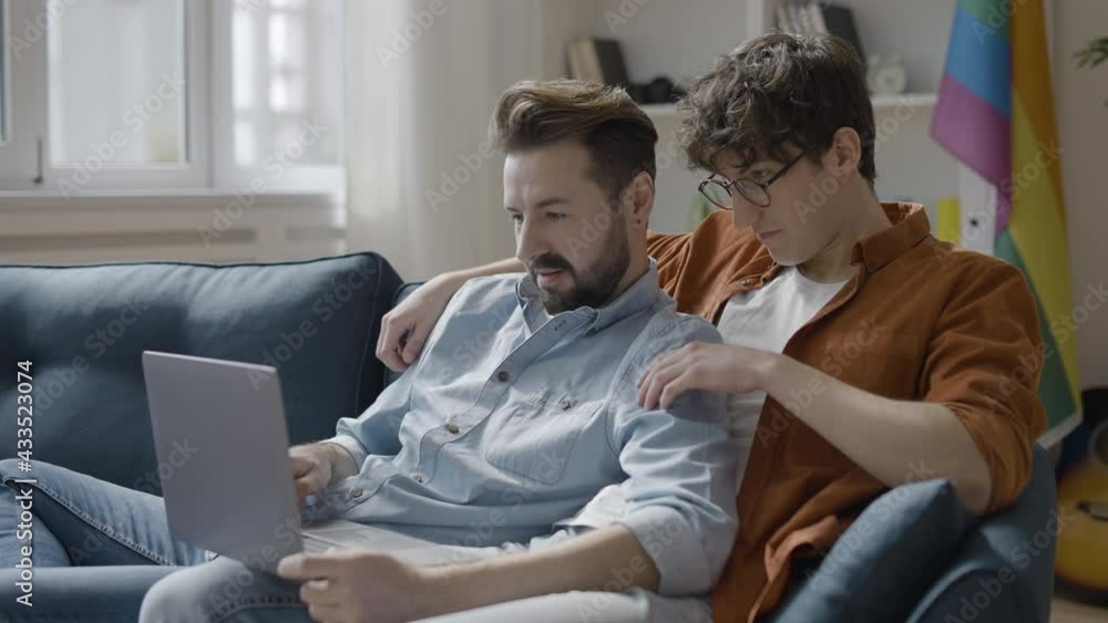 Homosexual couple sitting on couch, looking at laptop screen, booking services Homosexual couple sitting on couch, looking at laptop screen, booking services