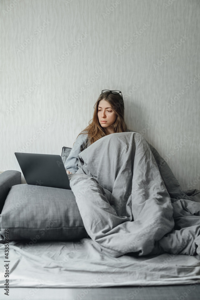 Sick girl with fever and flu sits in bed. Looks at the laptop. Sick girl with fever and flu sits in bed. Looks at the laptop.