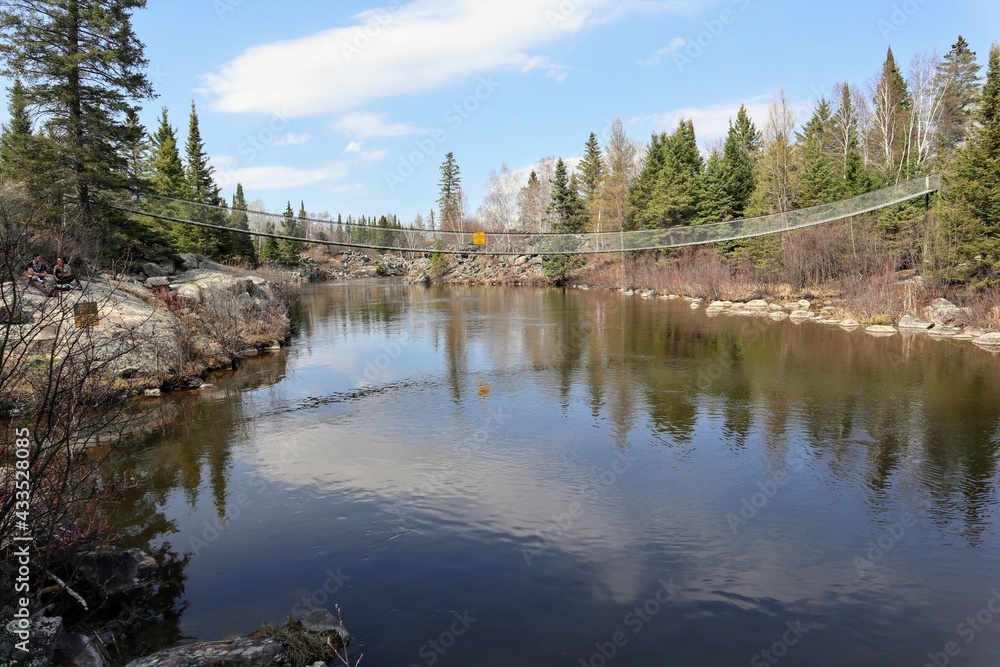 Suspension bridge over calm, narrow river flowing between high banks, overgrown with forest. Suspension bridge over calm, narrow river flowing between high banks, overgrown with forest.