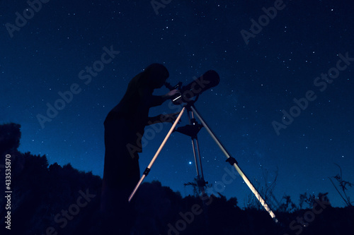 Dark silhouette of a sky watcher of the starry sky in a telescope. Woman observer watching night sky in a reflector. Image may contain noise grain due to high ISO range when shooting very dark scenes.