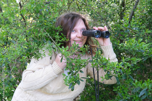 sly curious European adult woman holding black field binoculars with zoom in her hands, hiding in greenery, peeping out of green bushes, spying on unfaithful husband, neighbors, nature observation photo