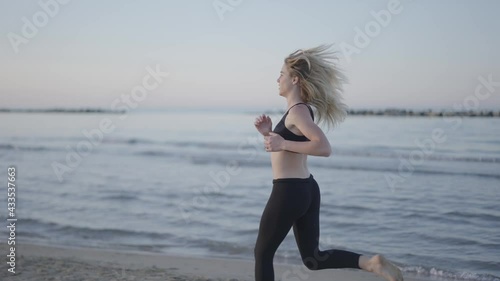 Steady shot of a pretty young woman running at the beach along the shore by the ocean at sunset. Girl works out for wellbeing in summer.   photo