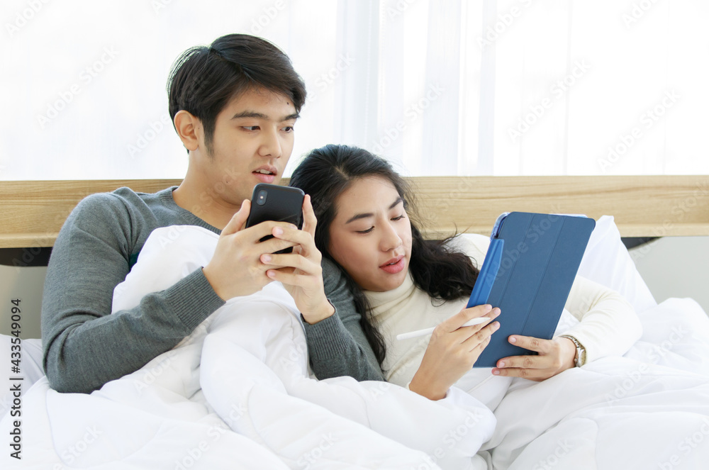Portrait of cute smiling young Asian lover couple in white and gray long sleeve sweatshirt lying on a bed while using a digital tablet together with a woman holding white digital stylus pen Portrait of cute smiling young Asian lover couple in white and gray long sleeve sweatshirt lying on a bed while using a digital tablet together with a woman holding white digital stylus pen