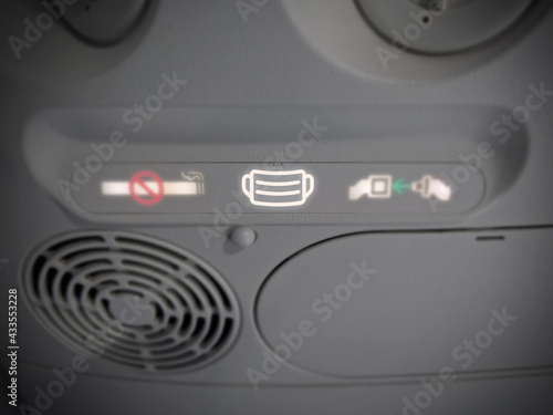 Airplane mask wearing mandate icon symbol for COVID-19 pandemic next to no smoking and fasten seat belt sign on overhead passenger service unit (PSU). Facial coverings are required on airline flights.