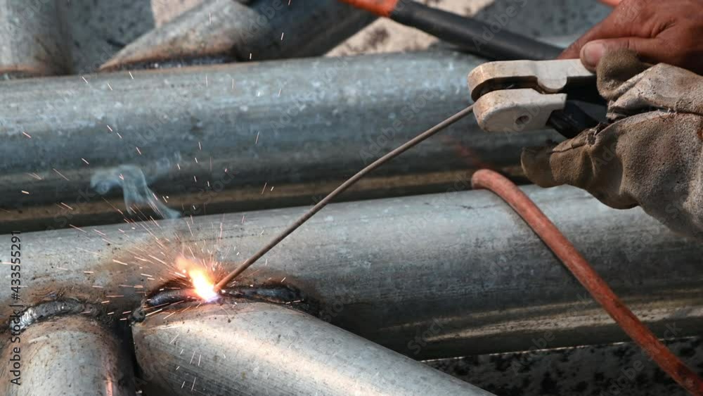 A welder is welding a steel pipe and is wearing a mask and leather gloves.