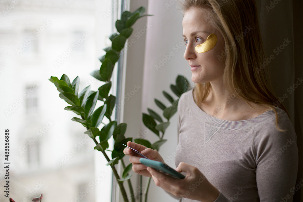 A European woman makes online purchases standing by the window and taking care of her skin. A European woman makes online purchases standing by the window and taking care of her skin.
