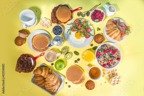 Various traditional breakfast food - fried eggs with bacon, muesli, oats, waffles, pancakes, burger, croissants, fruit berry, coffee, tea and orange juice, yellow table background copy space top view