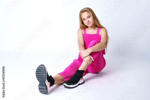 Young cute girl with long blond hair of Caucasian appearance posing in a pink suit on a white background sitting. Stylish and fashionable clothes for a teenager