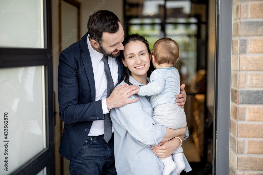 Happy husband hugs wife with one year old baby boy while standing in front of their front door house. Caucasian young family with baby. Happy husband hugs wife with one year old baby boy while standing in front of their front door house. Caucasian young family with baby.