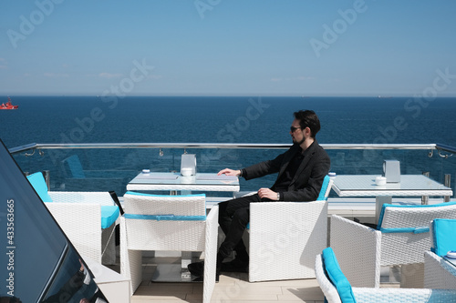 handsome young man in a suit sits on the terrace of a restaurant by the ocean. a man sitting at a table in a bar on a passenger cruise ship © Irina