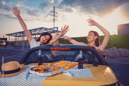Young girls smiling, raised up hands, posing in yellow car with french fries, pizza and soda in glass bottles on its trunk. Fast food. Copy space © nazarovsergey