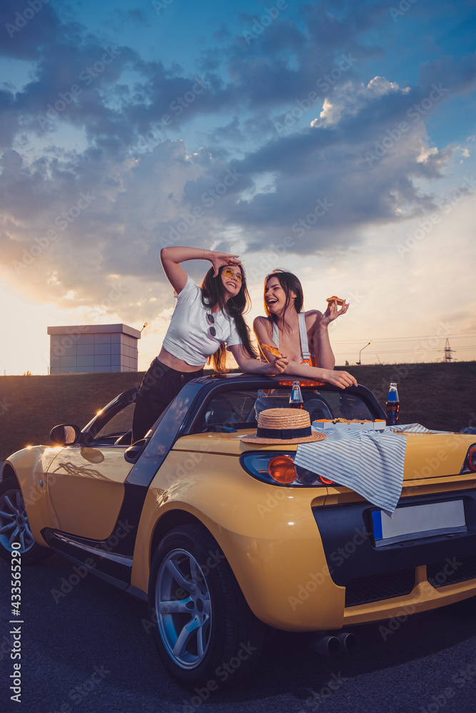 Two young models eating pizza, laughing, posing in yellow car cabriolet with hat and soda in glass bottles on its trunk. Fast food. Copy space