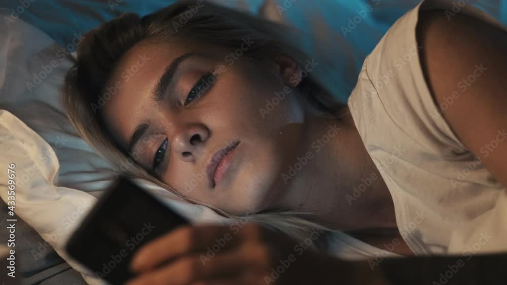 Gadget night. Late home leisure. Insomnia problem. Internet addiction. Tired sleepy woman with calm face reading online using mobile phone lying in cozy bed in dark bedroom. Gadget night. Late home leisure. Insomnia problem. Internet addiction. Tired sleepy woman with calm face reading online using mobile phone lying in cozy bed in dark bedroom.