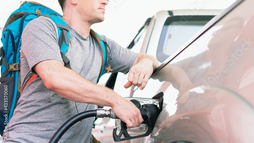 White caucasian car owner with blue backpack holding black fuel nozzle and filling high energy power fuel in auto car tank in petrol station, commercial service for benzine, diesel, gasohol, gasoline