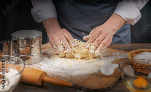 Women's hands, flour and dough. A woman in an apron cooking dough for homemade baking, a rustic home cozy atmosphere, a dark background with unusual lighting.