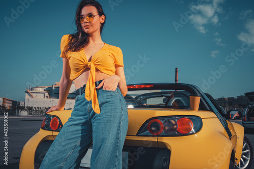 Girl in blue jeans, orange top and sunglasses is posing leaning on yellow car cabriolet with paper cup of beverage on trunk at parking lot. Copy space photo