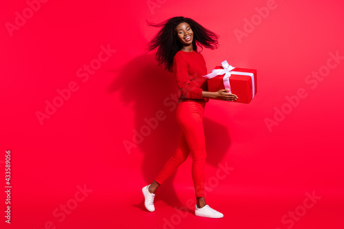 Photo of impressed cute dark skin lady wear casual sweater walking holding present box smiling isolated red color background