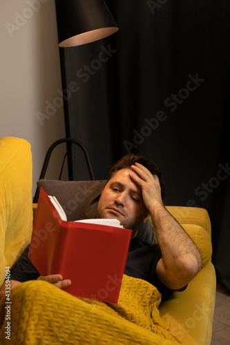 Young unshaven handsome guy reads book liyng on comfortable yellow sofa. male reader took his hand to head, reading detective or historic novel at home comfy couch covered with plaid blanket