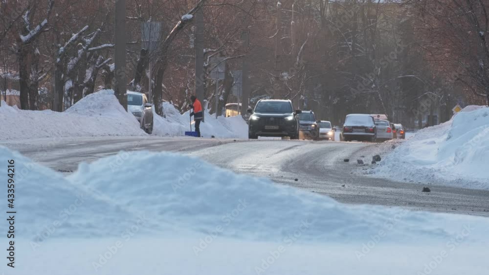 Automobiles drive along road with high snowdrifts on roadsides and working employee in orange vest on city street on winter day Automobiles drive along road with high snowdrifts on roadsides and working employee in orange vest on city street on winter day