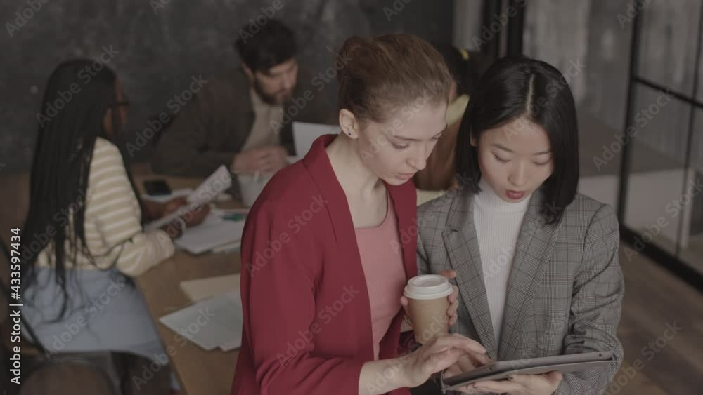 High angle medium close-up of young female Asian and Caucasian colleagues wearing formal attire, standing in conference room in foreground of colleagues working at table, talking High angle medium close-up of young female Asian and Caucasian colleagues wearing formal attire, standing in conference room in foreground of colleagues working at table, talking