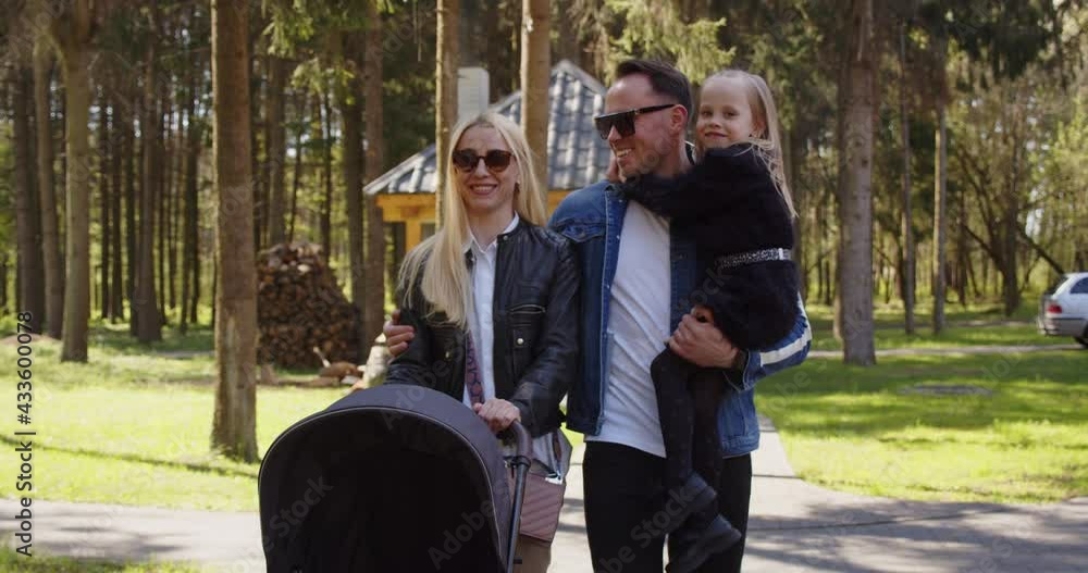 Happy mom dad and older child are walking along park and rolling before them stroller with small baby and smiling. Happy mom dad and older child are walking along park and rolling before them stroller with small baby and smiling.