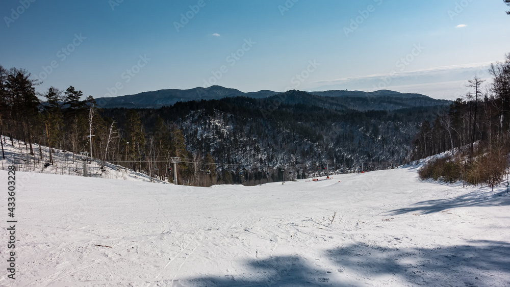 A chairlift runs over the snow-covered mountain slope. Ski tracks in the snow. Wooded mountain range against the background of a clear sky. Winter day