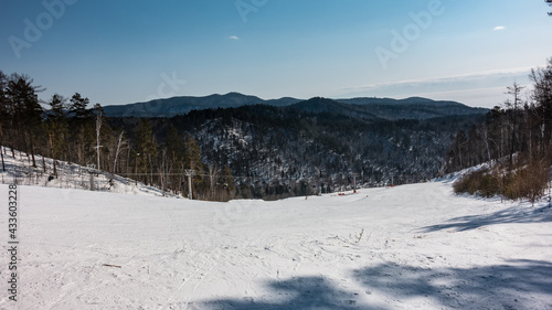 A chairlift runs over the snow-covered mountain slope. Ski tracks in the snow. Wooded mountain range against the background of a clear sky. Winter day